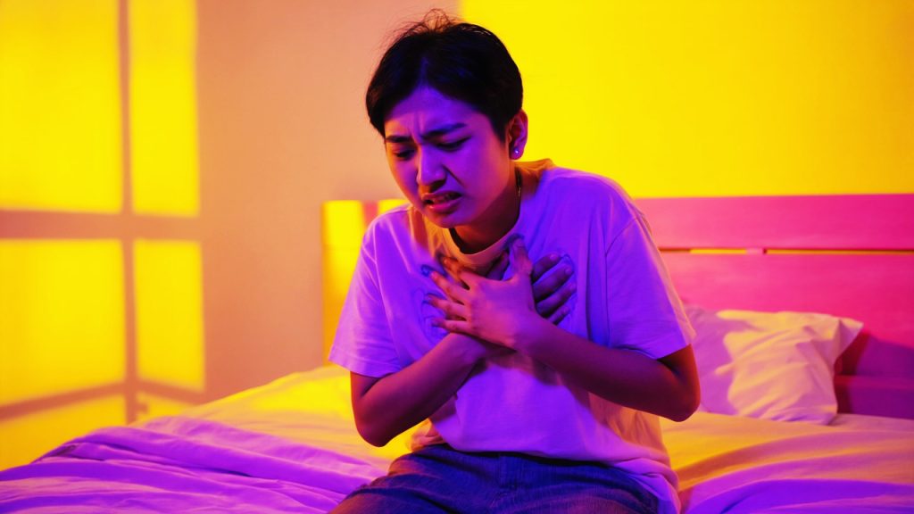 Uma pessoa sentada em um quarto, segurando o peito com uma expressão de medo e desorientação, simbolizando o início de um ataque de pânico e a sensação de terror avassalador.