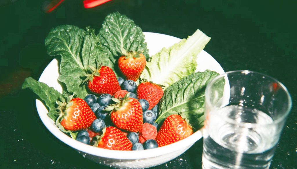 Tigela vibrante e colorida com frutas e folhas verdes e um copo de água, simbolizando a nutrição saudável como combustível essencial para a clareza mental e emocional.